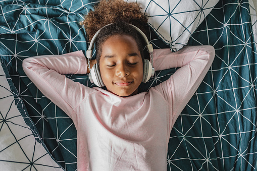 Young girl laying in bed enjoying music, she is listening to music via wireless headphones
