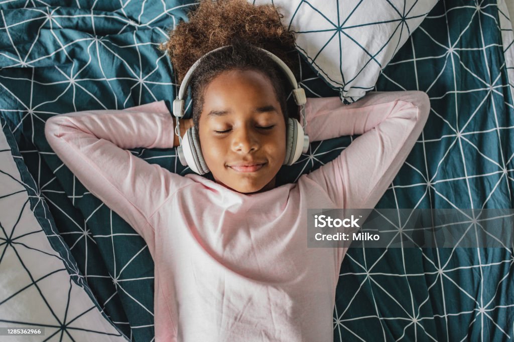 A little African American girl enjoys her free time at home Young girl laying in bed enjoying music, she is listening to music via wireless headphones Child Stock Photo