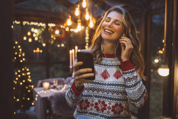 Portrait of happy modern young woman enjoying listening to music with headphones holding phone, looking away, wear hat, jacket in winter season, in forest against Christmas tree background