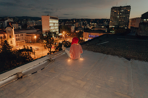 Photo of a young woman enjoying the city lights on a cold, winter night; celebrating holidays alone, on a rooftop of the building.