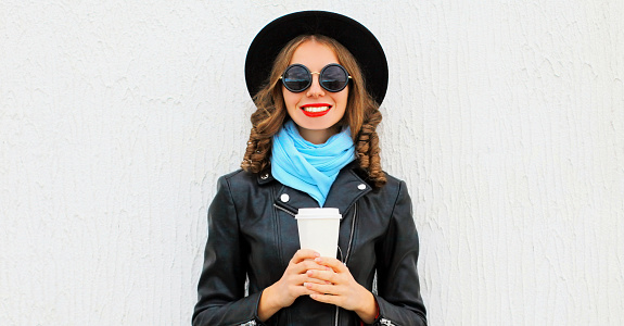 Portrait of beautiful smiling young woman with coffee cup wearing a black round hat, jacket, sunglasses in the city
