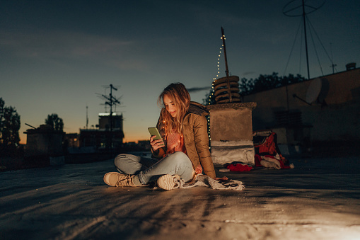 Photo of a young woman chatting online and checking her social media while enjoying her evening on a rooftop of the building.