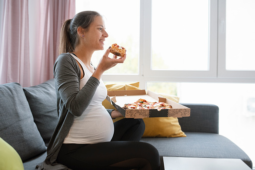 Side view of pregnant woman eating pizza at home.