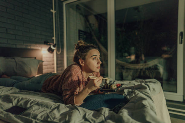 Binge watching my favorite show Photo of a young woman watching TV in the bedroom of her apartment; eating sushi and enjoying her night at home alone. watching tv stock pictures, royalty-free photos & images