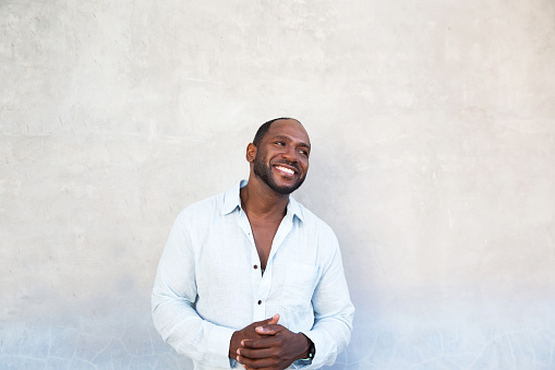 Handsome cheerful african american man in white shirt standing with grey blue textured wall. Casual style.