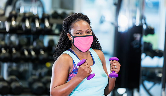 A mature African-American woman in her 40s working out at the gym, lifting hand weights. She is exercising during the covid-19 pandemic, wearing a protective face mask to prevent the spread of coronavirus.