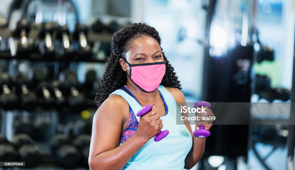 African-American woman at the gym, wearing face mask A mature African-American woman in her 40s working out at the gym, lifting hand weights. She is exercising during the covid-19 pandemic, wearing a protective face mask to prevent the spread of coronavirus. Gym Stock Photo