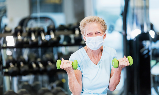 A senior woman in her 60s working out at the gym, lifting hand weights. She is exercising during the covid-19 pandemic, wearing a protective face mask to prevent the spread of coronavirus.