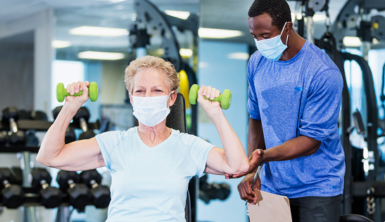A senior woman in her 60s working out at the gym, lifting hand weights. Her personal trainer, an African-American man in his 30s, is watching her, adjusting her arm position.. She is exercising during the covid-19 pandemic. They are wearing protective face masks to prevent the spread of coronavirus.