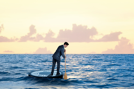 A businessman stands on a floating compass as he paddles out to the open sea using a wooden paddle in search of his destination. The compass is surrounded by a blue sea as a late afternoon sun glints off the surface as he realizes his insignificance in a large and dangerous world.