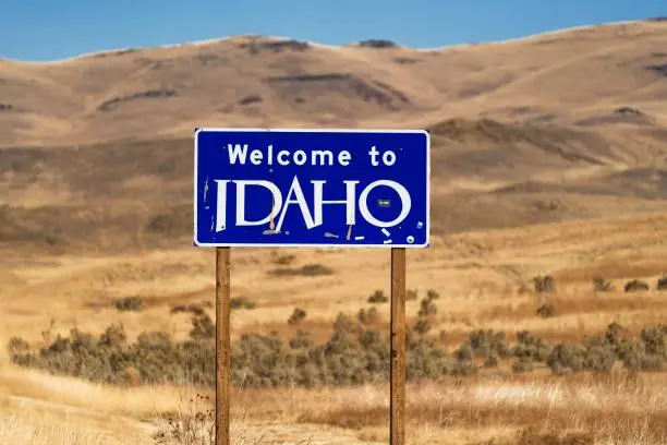 This image shows an official "Welcome To Idaho" state road side sign with a beautiful landscape behind.