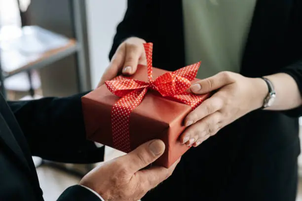 Close-up view of hands of unrecognizable woman giving red gift box tied to bow handed to man. Giving gifts during the Christmas, Happy New Year and Happy Birthday at office.