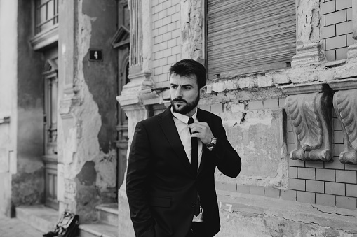 Portrait of a handsome stylish male outdoors, walking. Having a strong facial expression while looking on the right, placing the tie on its place with one hand in the pocket. Black and white retro concept.