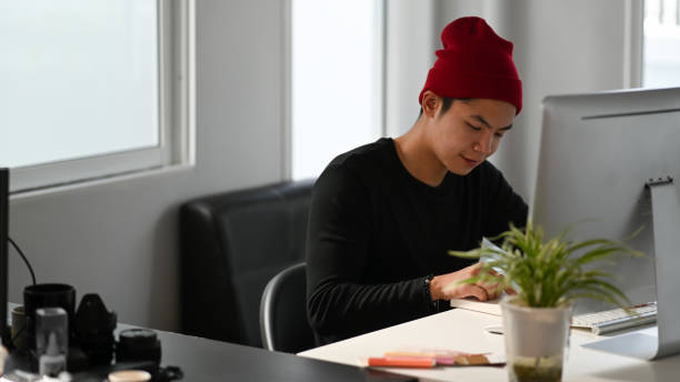 A male creative graphic designer is reading a book while sitting in front of computer at workplace.