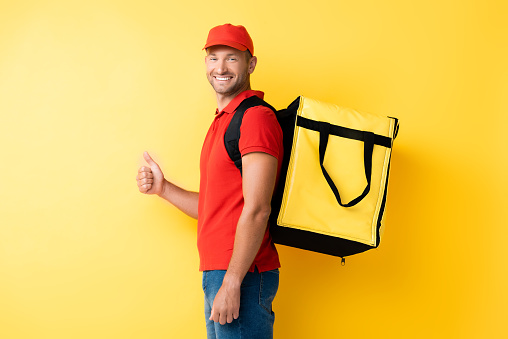 cheerful delivery man carrying backpack with order and showing thumb up on yellow