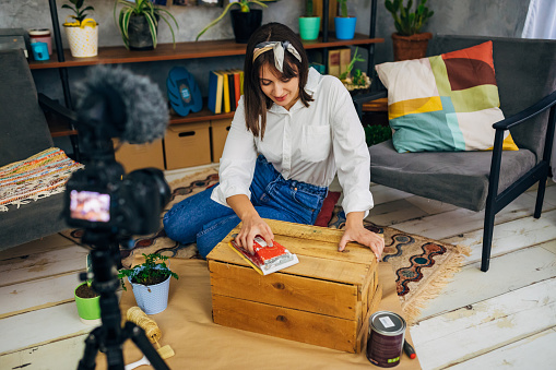 Female vlogger recording her tutorial about wood sanding and easy DIY how to paint a furniture from your home, while using camera for vlogging in her cozy home during COVID-19 pandemic