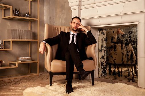 Young businessman man in suit is sitting on chair in living room holding hand on his head. Gesture of person indicates calmness, relaxation and home comfort. Concept of human emotions. Copy space
