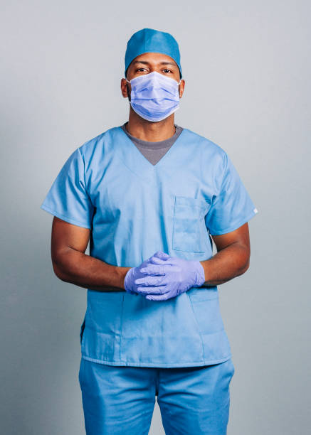 Male surgeon with hands clasped during COVID-19 Confident surgeon in blue scrubs during COVID-19. Portrait of male nurse wearing protective workwear during pandemic. He is standing against gray background. medical scrubs stock pictures, royalty-free photos & images