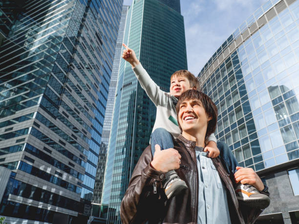 istock Little boy sits on father's shoulders and points on skyscraper. Dad and son looks on glass walls of buildings. Future and modern technologies, life balance and family life in well keeps districts. 1285441296
