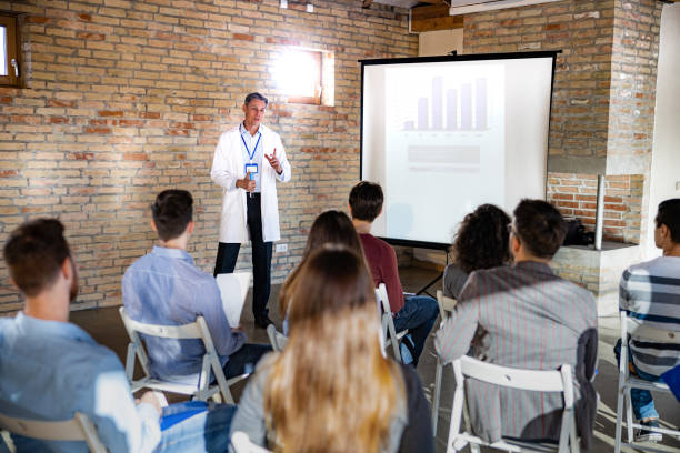 Male doctor talking to group of people on presentation in the hospital. Mid adult male doctor talking to large group of people during a presentation in board room. medical education stock pictures, royalty-free photos & images