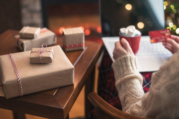 istock Woman holding credit card using laptop for making order sitting at table with packaging gift, cup of cocoa and marshmallows near fireplace and christmas tree. Online shopping concept 1285445349