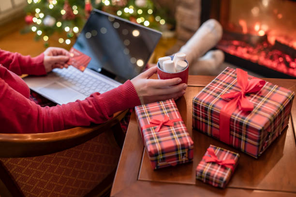 istock Woman holding credit card using laptop for making order sitting at table with packaging gift, cup of cocoa and marshmallows near fireplace and christmas tree. Online shopping concept 1285446702