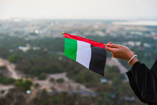 Emirati woman wearing hijab holding United Arab Emirates flag in the desert mountain at sunset