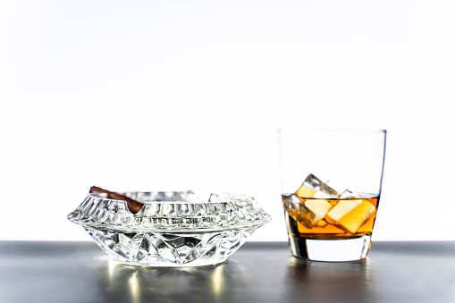 Glass of whiskey with ice cubes and cigar in crystal ashtray on white background. Alcohol drink concept, copy space provided. The scene is situated in front white background in controlled studio environment . The photo is taken with Sony A7III camera.