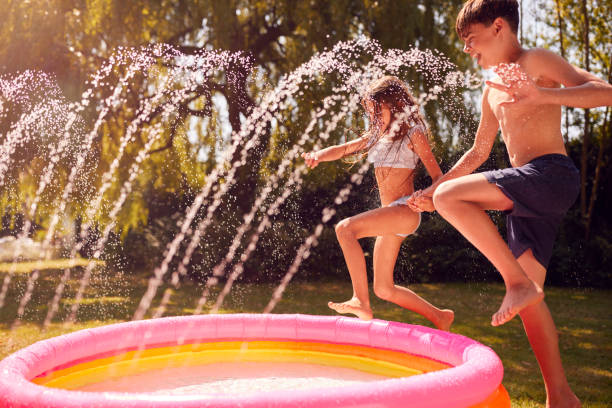 Children Wearing Swimming Costumes Having Fun In Garden Playing In Water From Garden Sprinkler Children Wearing Swimming Costumes Having Fun In Garden Playing In Water From Garden Sprinkler staycation stock pictures, royalty-free photos & images