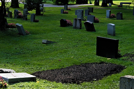 Cemetery with tilted headstones and dirt from a new grave on a green lawn.