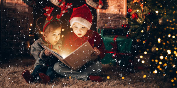Brother and sister are sitting and reading a book. Christmas mood. Children in New Year's costumes against the background of a Christmas tree and a fireplace stock photo