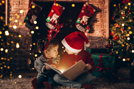 Brother and sister are sitting and reading a book. Christmas mood. Children in New Year's costumes against the background of a Christmas tree and a fireplace.