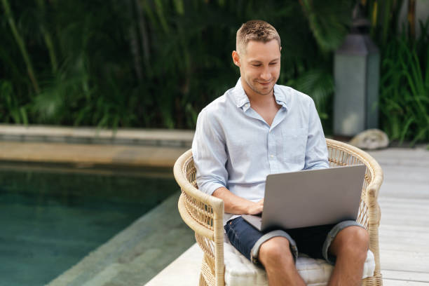 istock Young man using a laptop computer in a garden with a swimming pool. 1285455780