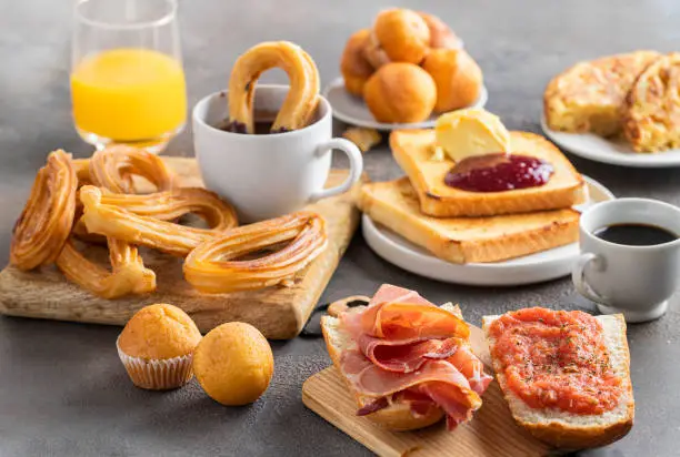 Different Spanish breakfast, tortilla, bruschetta with jamon (cured ham) and tomatoes, churros with chocolate,  Magdalena (Spanish cupcake) , crusty  french toast with butter and jam ,coffee and orange juice.