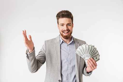 Portrait of an excited young man dressed in shirt and jacket holding money banknotes and celebrating isolated over white background