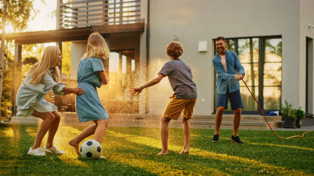 ガーデンウォーターホースで遊ぶ4人の幸せな家族は、お互いにスプレーします。母、父、娘、息子は晴れた夏の日に牧歌的な郊外の家の裏庭の芝生でゲームを楽しんでいます - humor men laughing teenager ストックフォトと画像