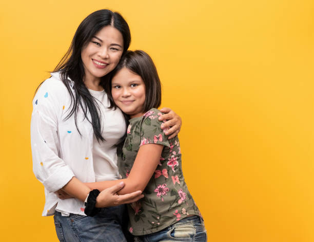 Happy family, smiling mom with her daughter. Young woman and teen girl dressed in casual clothes poses embracing in studio. Isolate on a yellow background. Copy space on right side Happy family, smiling mom with her daughter. Young woman and teen girl dressed in casual clothes poses embracing in studio. Isolate on a yellow background. Copy space on right side. adoption photos stock pictures, royalty-free photos & images
