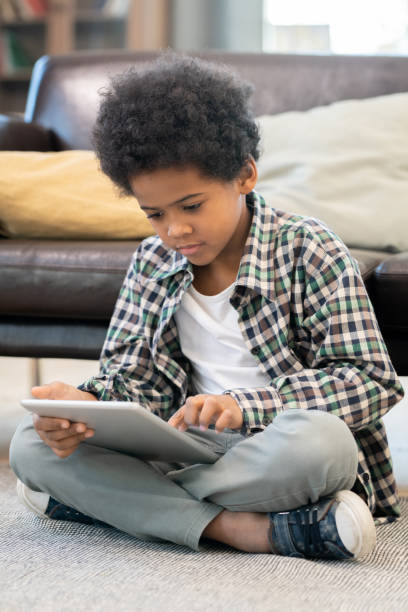 istock Serious elementary schoolboy of African ethnicity sitting by black leather couch 1285467180