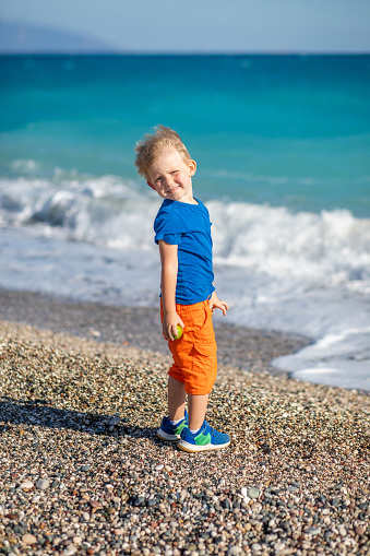 Smiling beautiful blond child stands on the beach of the blue and turquoise sea of the ocean. The wind blows his flowing hair. Bright vertical shot concept about childhood and happiness, lifestyle.