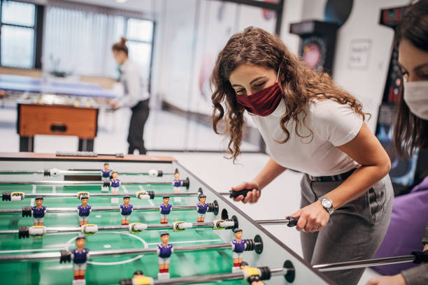Female colleagues playing foosball in firm's playroom Group of women colleagues playing foosball on a break from office work, in firm's playroom. They are all wearing protective mask to protect from coronavirus spreading. woman darts stock pictures, royalty-free photos & images