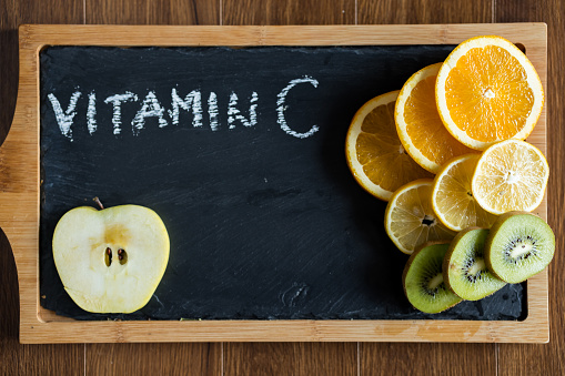 Orange, kiwi, apple and lemon slices with words vitamin c written on black cutting board.