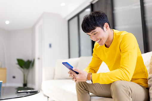 Asian young man uses smartphone happily in the living room at home