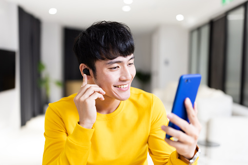 asian young man wearing earbud is having video chat or listening podcast on smartphone with his friend at home