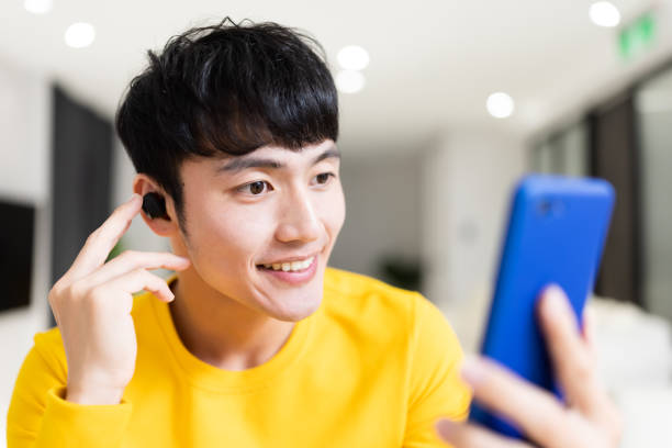 asian man has video chat close up of asian young man wearing earbud is having video chat or listening podcast on smartphone with his friend at home in ear headphones stock pictures, royalty-free photos & images
