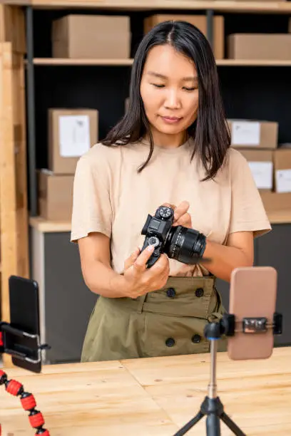 Asian young woman in casualwear holding photocamera over table while unpacking it and recording the process on smartphone camera