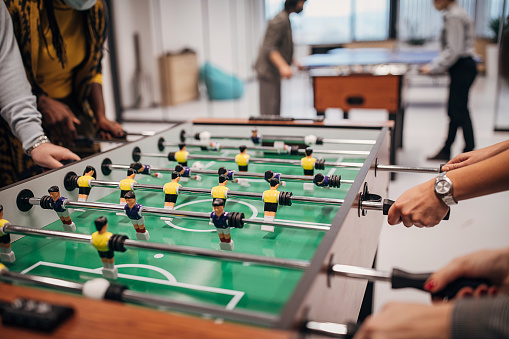 Group of people, man and women colleagues playing foosball on a break from office work, in firm's playroom. They are all wearing protective mask to protect from coronavirus spreading.