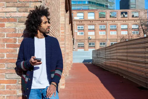 Photo of Young handsome African man using his smartphone with smile while leaning on a bricked wall outdoors in sunny day