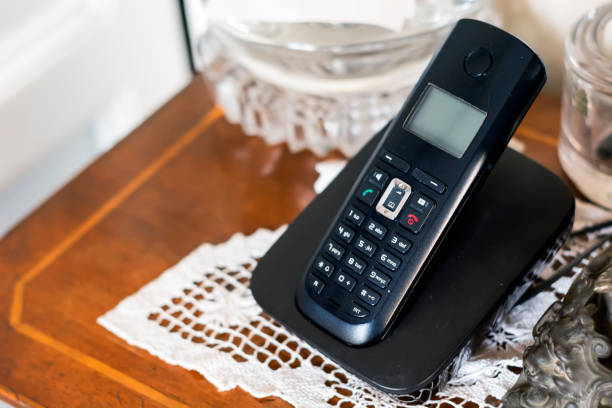 An old cordless telephone on its charging cradle a black plastic cordless phone charging on its cradle inside a classically furnished home with antiques and white crochet. Outdated technology landline phone stock pictures, royalty-free photos & images