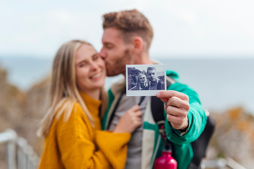 A young caucasian couple are standing by the sea in Polperro, Cornwall. The male is kissing his girlfriend on the cheek whilst holding up a polaroid instant photo they just took together. The focus is on the polaroid in the foreground.