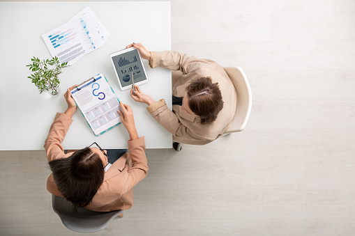 Overview of two young elegant businesswomen sitting by desk, analyzing financial charts and diagrams and discussing the data at meeting